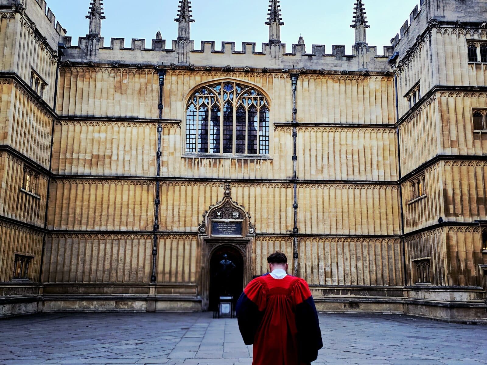 Peter McCann Strain in scarlet Oxford doctoral robes walking toward the lit gothic doorway of the Bodleian Library quad at dusk